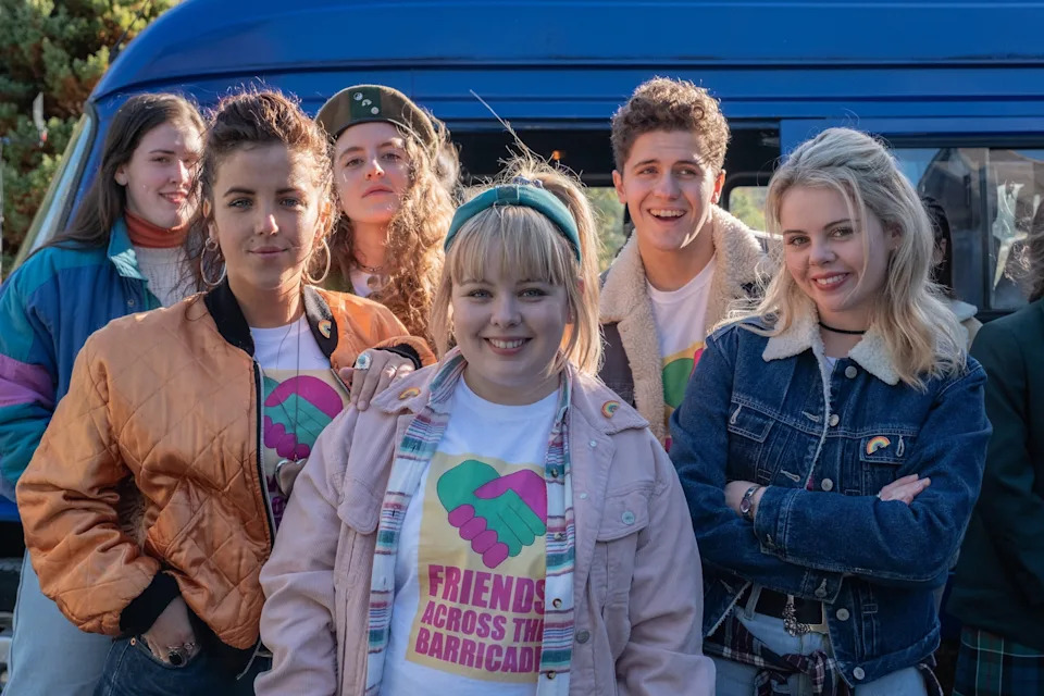 Group of young people in casual 90s style clothing stand smiling by a van. They wear shirts that read "Friends Across the Barricades."
