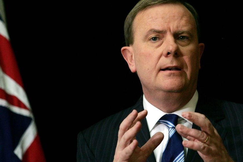 Peter Costello wearing and suit and gesturing with his hands, with an Australian flag nearby.