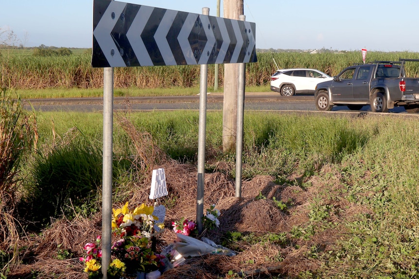 A road sign with flowers and notes at road intersection with two cars in the background.