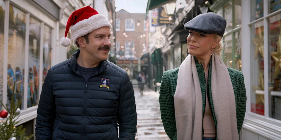 Two people walk down a festive street: one in a Santa hat and casual jacket, the other in a newsboy cap, blazer, and scarf