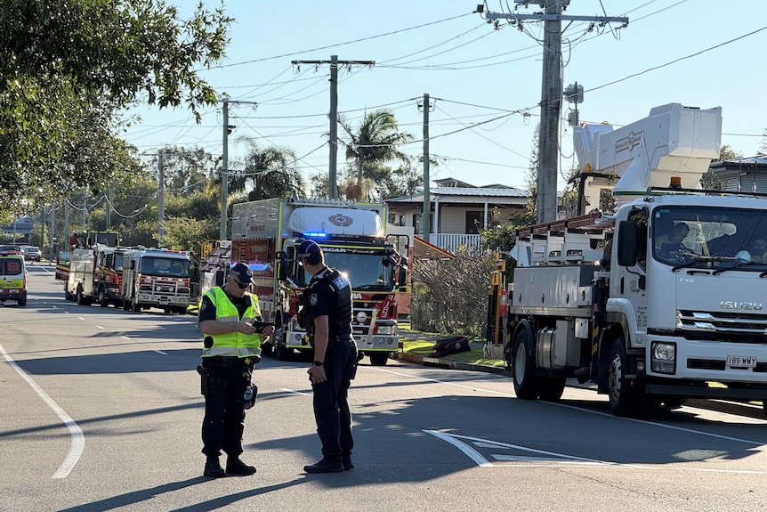 Two police officers stand on the road with fire trucks parked at the side.