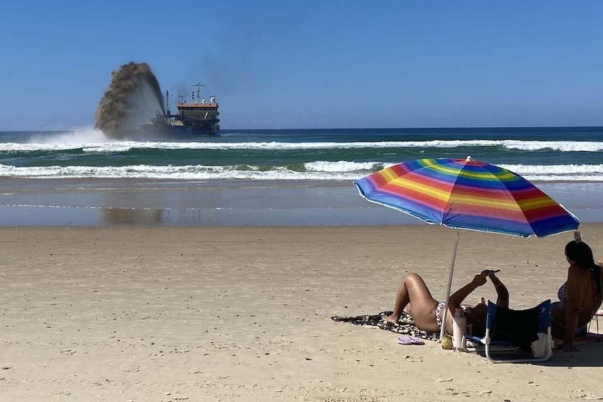 People relaxing on the beach as a barge pumps sand in the distance.
