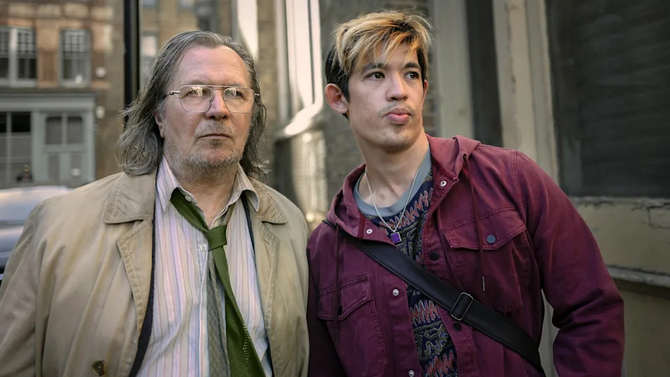 Two individuals stand together on a city street; one is older with long hair and a trench coat, the other younger with highlighted hair and casual attire
