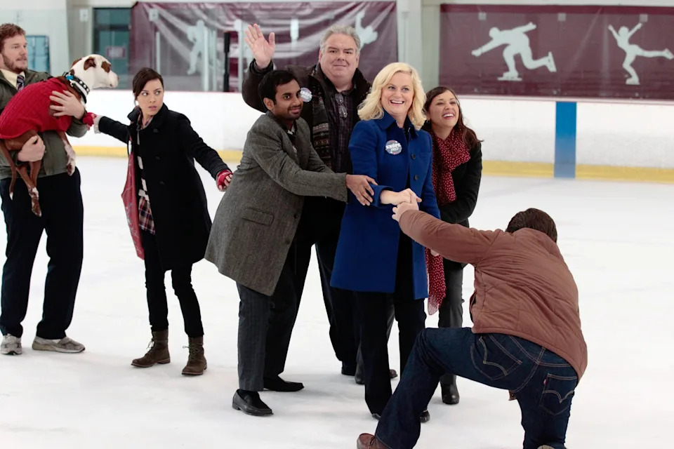 Group of people in winter attire having fun on an ice rink. A man kneels playfully in front of a woman in a blue coat