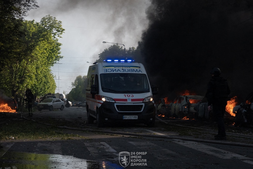 An ambulance drives down a road  lined with burning vehicles.
