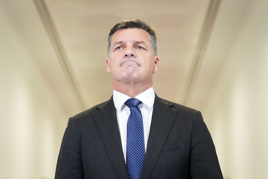A man with dark hair that is greying at the temples wears a dark suit as he stands in a corridor.