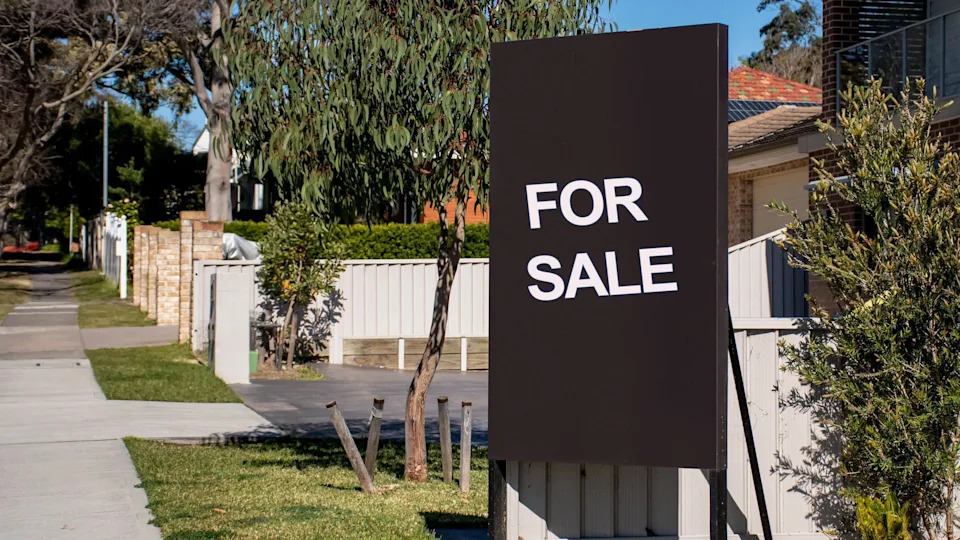 A "For Sale" sign on a suburban street with with houses and gardens behind