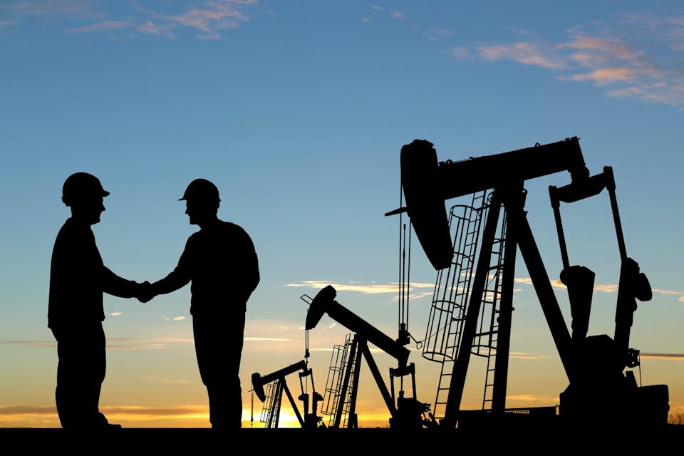 Two oil company employees shake hands in front of an oil production field.