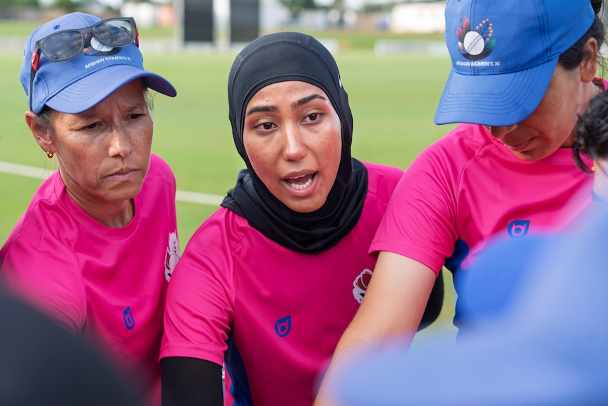 Three women in view, one in centre mid-speech, wearing a pink jersey, black hijab, all standing on a green sports field.