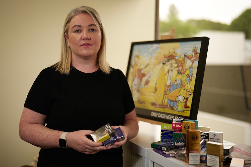 A blonde woman wears a neutral facial expression, in her hands she is holding cigarette packets.