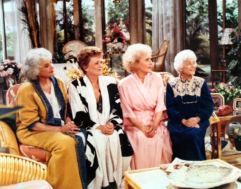 Four women in a living room set, wearing loungewear and robes, reminiscent of a classic TV sitcom scene