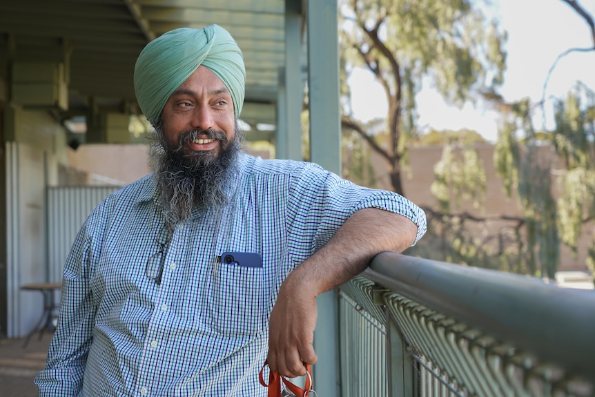 A man in a checked shirt and turban stands outside with his elbow leaning on a balcony railing starting off into the trees.