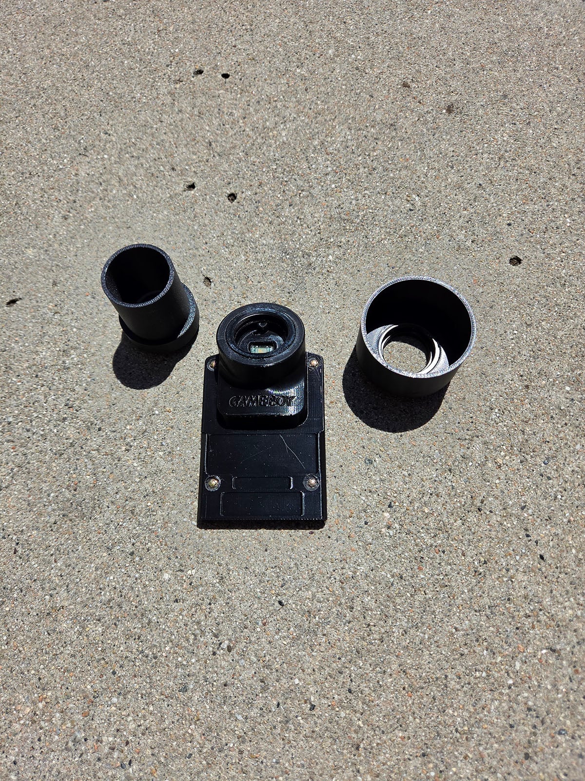 Three camera lenses and a black metal mount are arranged on a textured concrete surface in bright sunlight. The mount is in the center with a lens attached, and two separate lenses are placed on either side.