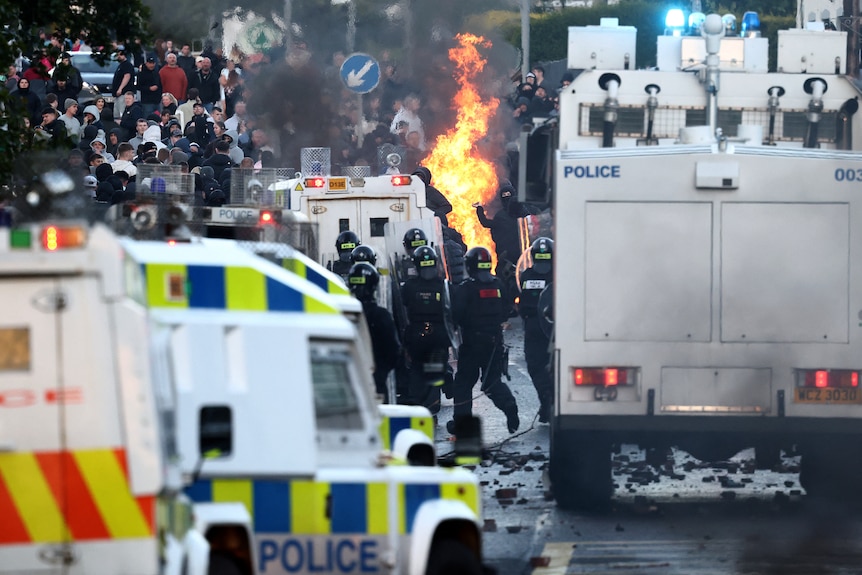 Police in black riot gear with white police vans confront a mass of people throwing projectiles in front of a fire.