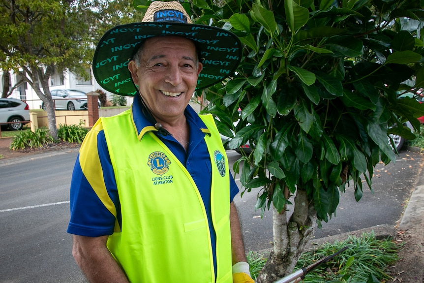 Man wearing yellow high-vis vest and large hat holding garden shears standing next to tree