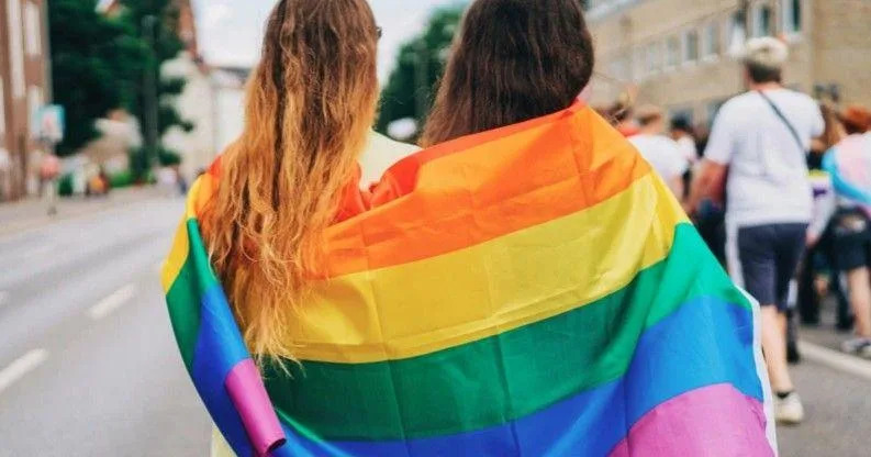Two people stand with their back to the camera, wrapped in a rainbow flag as a parade proceeds down a street