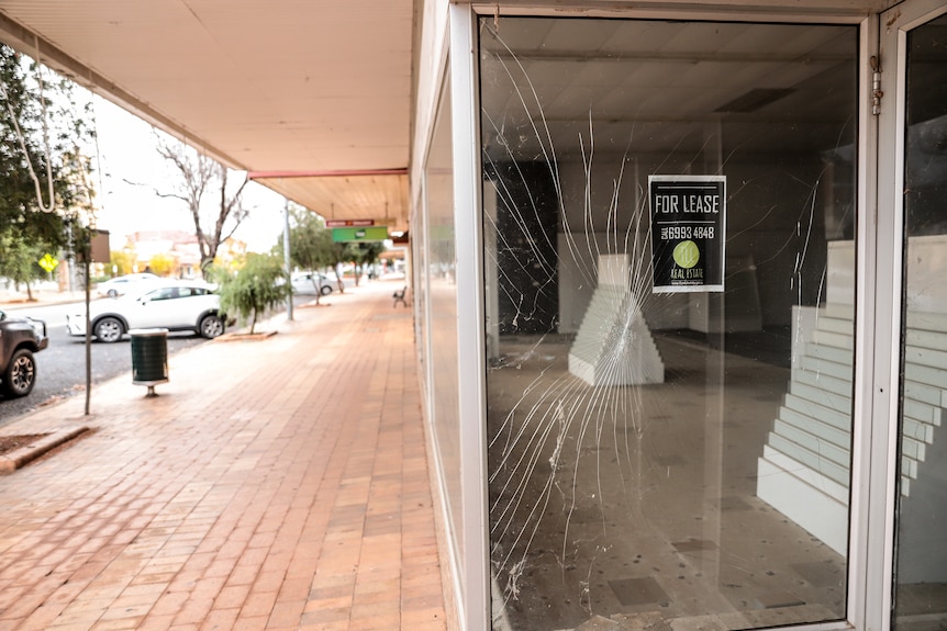 Shattered glass in the window of a shop next to a sign reading 'For Lease'. There is a footpath and parked cars to the left.