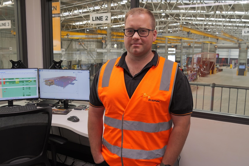 Man with glasses stands in front of warehouse in an organse high-vis vest.