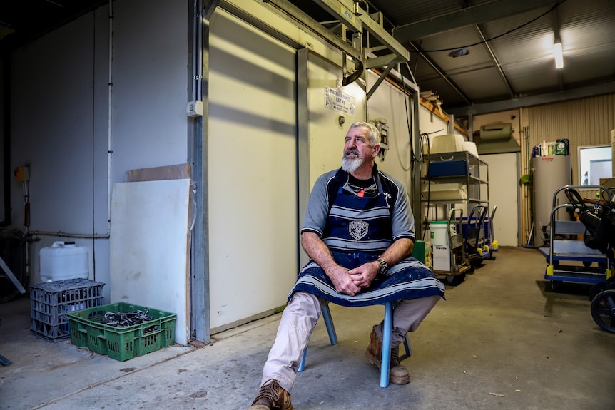 A man sits on a chair, wearing a blue and white striped butcher's apron. There is a cool room and equipment in the background.e