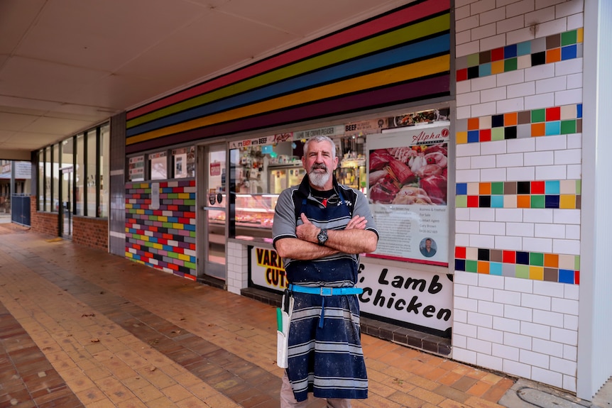 A man wearing a blue and white striped apron stands with his arms crossed outside a butcher shop with signs for meat behind him.