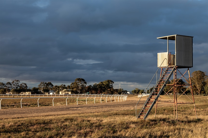 A tin box on a stand with a ladder up to it overlooks a dirt track and white railing of a race track. Clouds and trees behind.