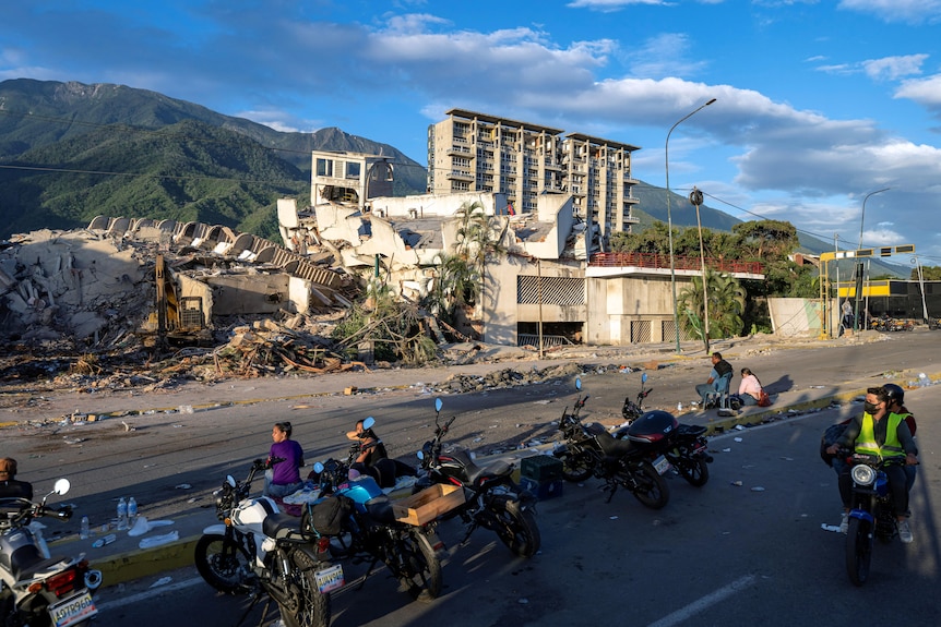 Piles of collapsed buildings alongside a dust-covered road and people standing alongside motorcycles.