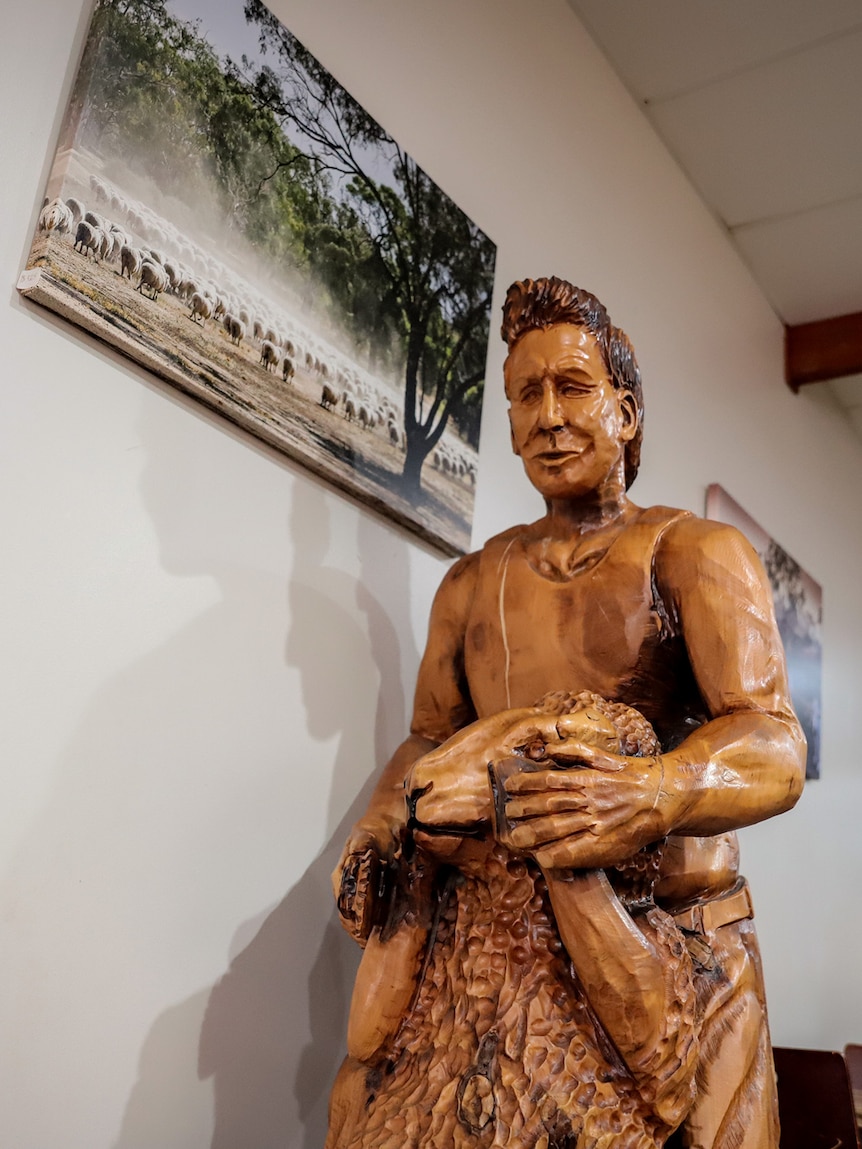 A carved wooden statue of a man wearing a singlet, holding a sheep is seen in a room with photos of farming scenes on the wall