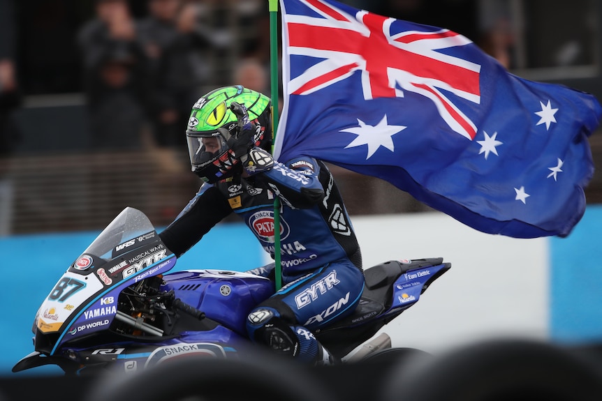 An Australian World Superbikes rider rides his bike carrying an Australian flag after a race win.