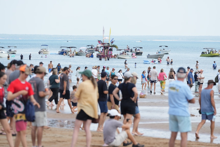 Crowd of people stand on in beach with tin boats in water ahead