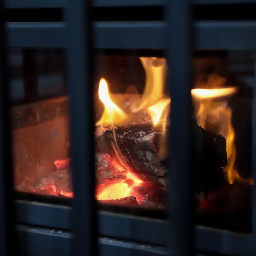 A burning log with embers and flames in a fireplace behind glass and metal grill bars.