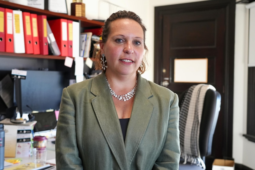 A dark-haired woman wearing a green blazer stands in front of a desk with a large computer monitor and many files on it.