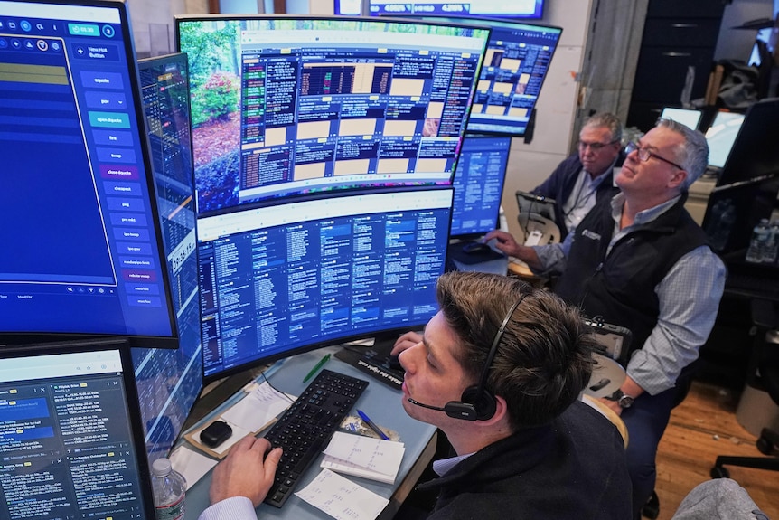 Male stock brokers surrounding multiple computer screens showing blue Wall Street charts.