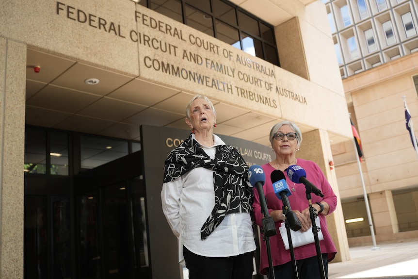 Two women with short grey hair stand in front of a microphone outside the federal court building.