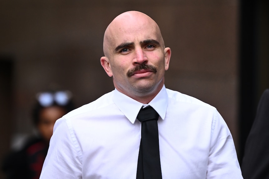 A man with a moustache in a white shirt and black tie walks outside court.