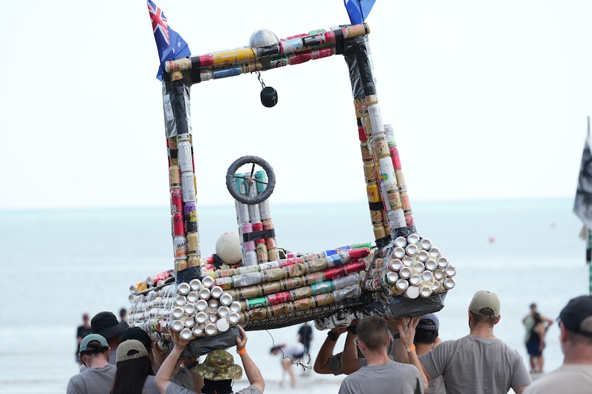 Group of people hold up boat made of beers cans above heads in front of water
