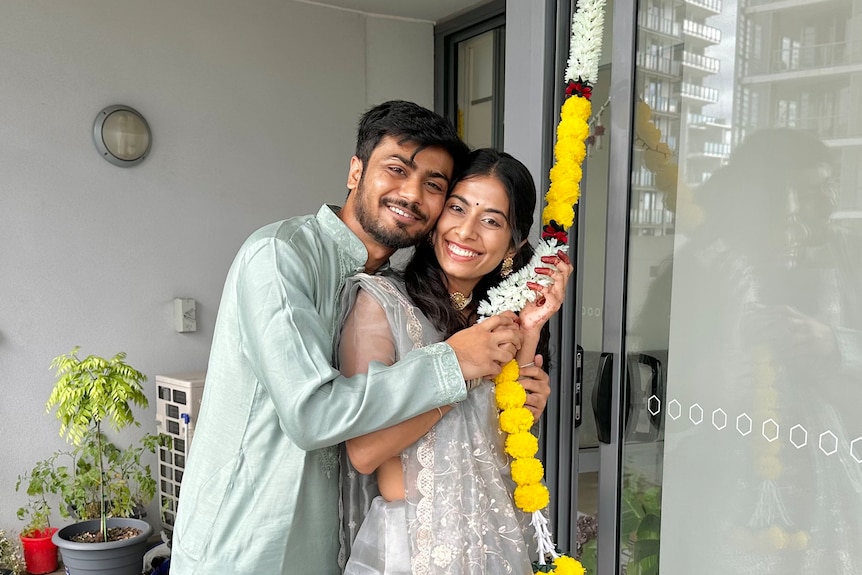 A young couple hold a string of flowers while smiling at the camera