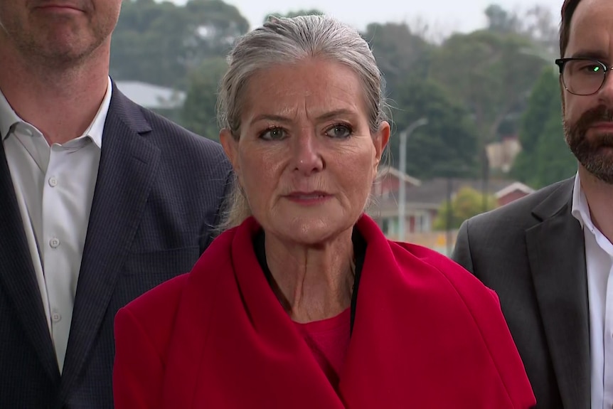 A woman with grey hair pulled back in a ponytail in a red coat stands in front of two men, staring ahead.