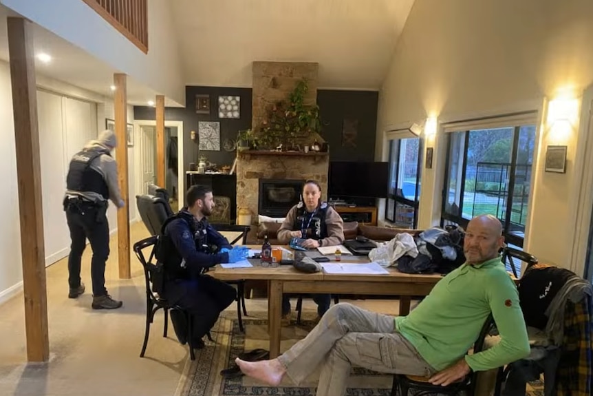 A police officer stands near two others who sit at a table with a bald man in a green shirt who look directly at the camera.