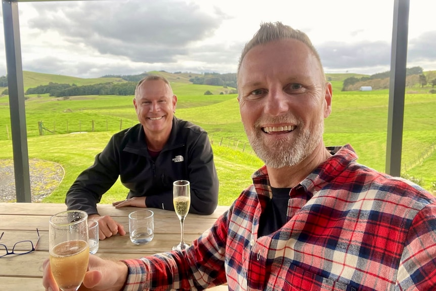 A man in a red patterned shirt and a man in a black jacket smile at the camera while at a wine tasting