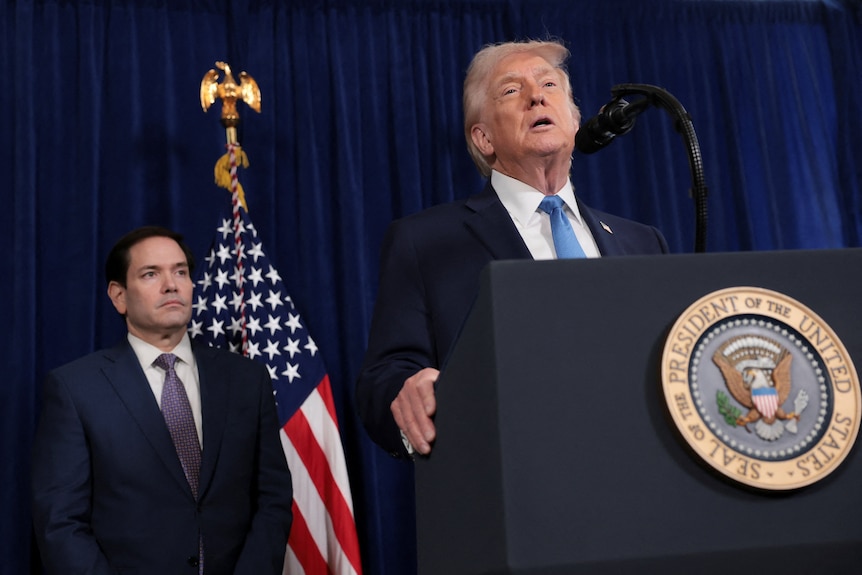 Donald Trump wearing a dark suit holds his hands up and speaks to reporters, Rubio stands behind.