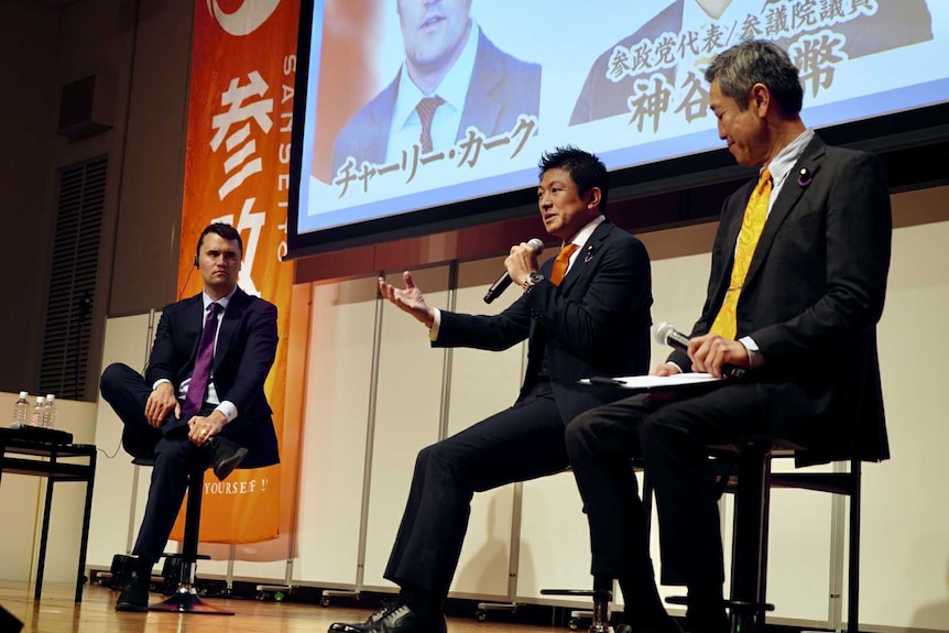 Charlie Kirk sitting next to Sanseito leader, Sohei Kamiya who is speaking into a microphone.