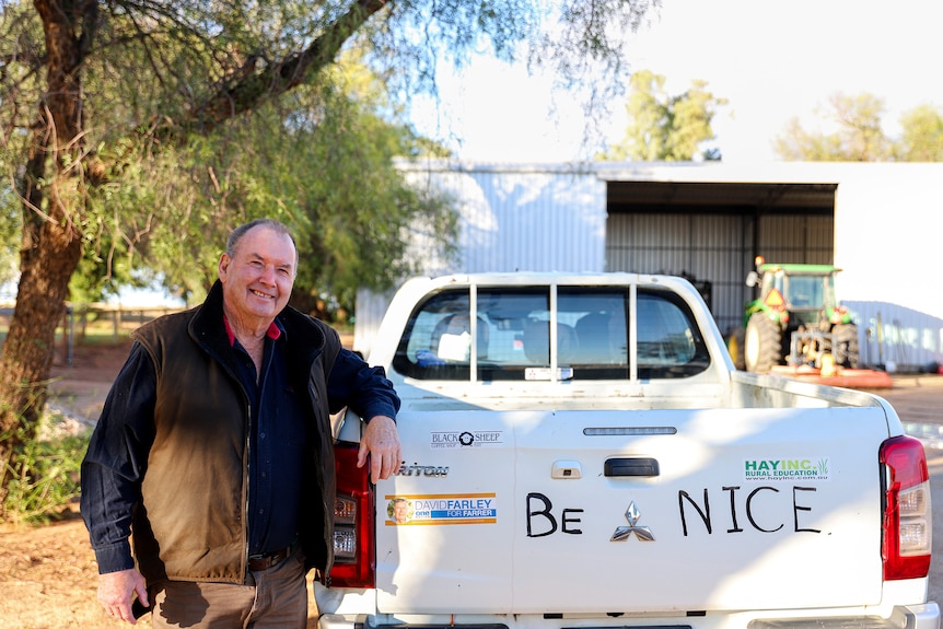 A man wearing a blue shirt and a brown jacket leans on a white utility vehicle that has writing saying 'Be Nice'