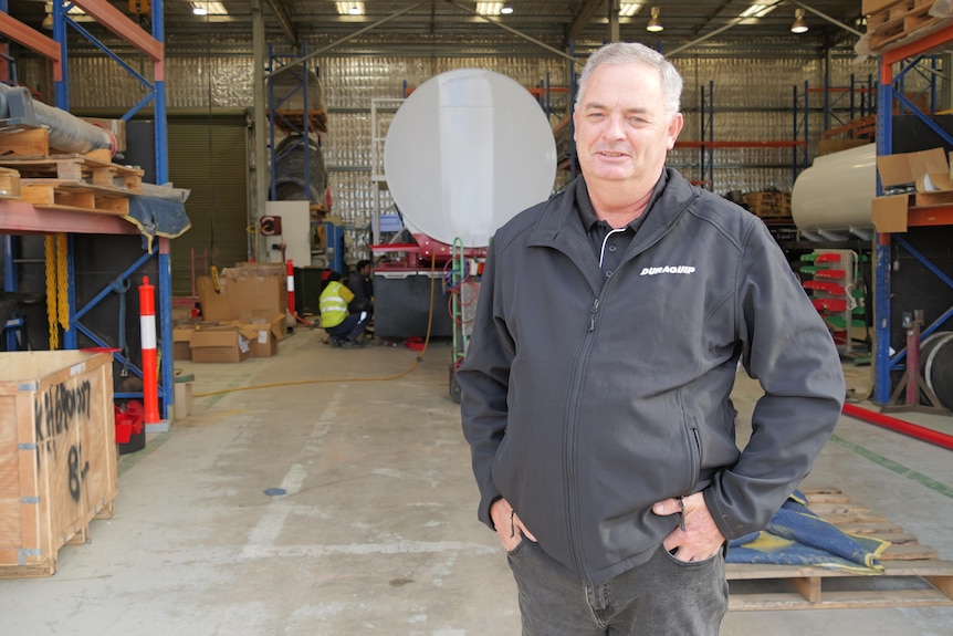 Middle-aged caucasian mean with grey hair stands in a warehouse with hands in the pockets of a black jacket.