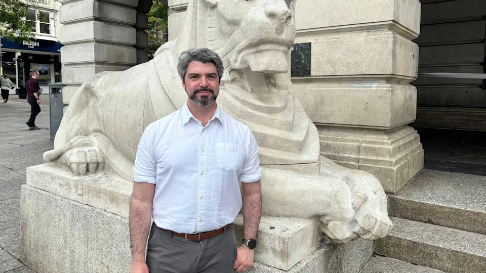 Man stood in front of stone lion statue. He is wearing a white shirt and smiling.