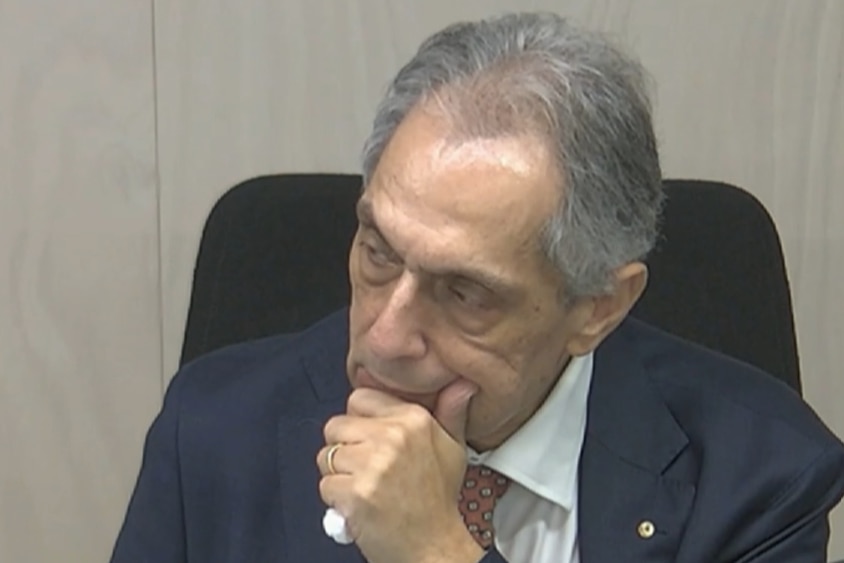 John strokes his chin and has a look of concentration. He has grey hair and a dark blazer.