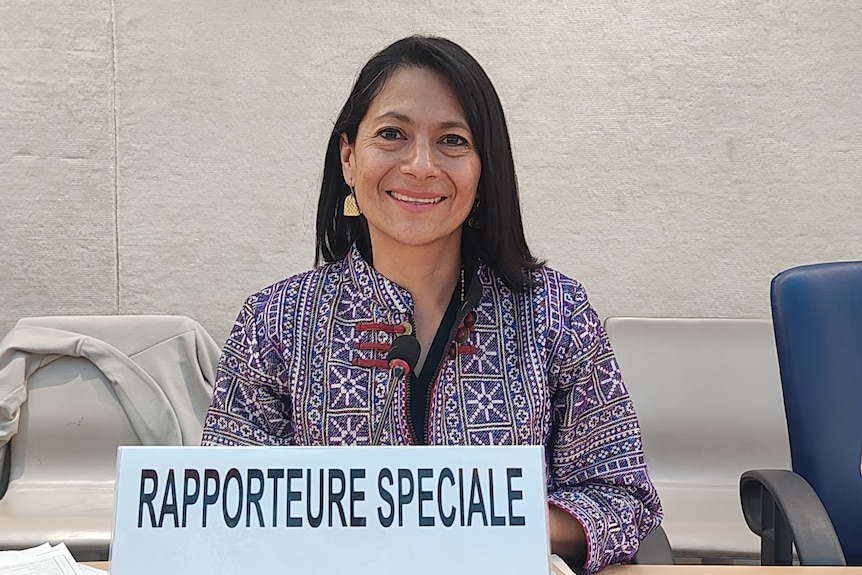 A Hispanic woman with black hair and wearing a colourful jacket sits before a placard reading "Rapporteure Speciale" in French.