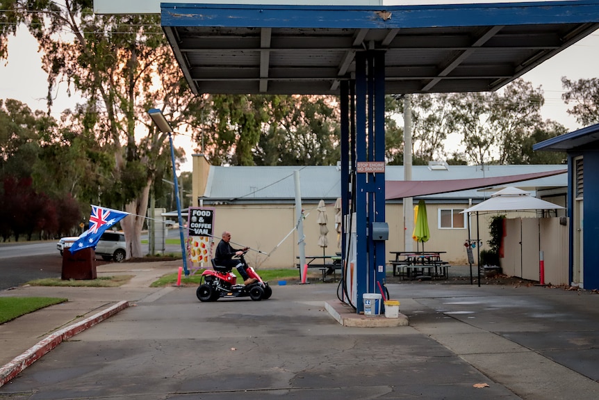 Man driving four wheel vehicle holds large Australian flag as he drives through country service station at dusk