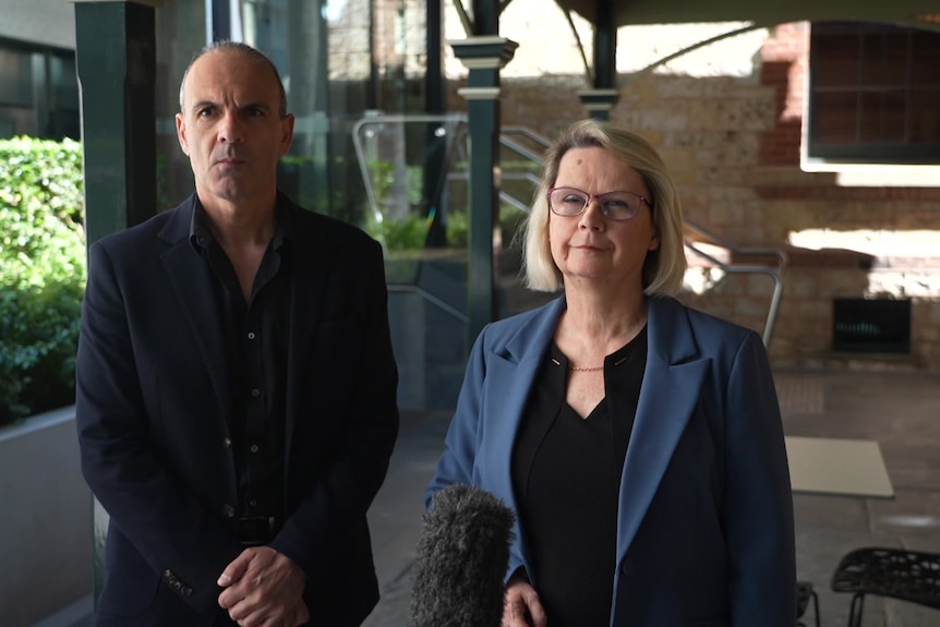 A man and a woman stand in front of microphones under a pergola next to hedges