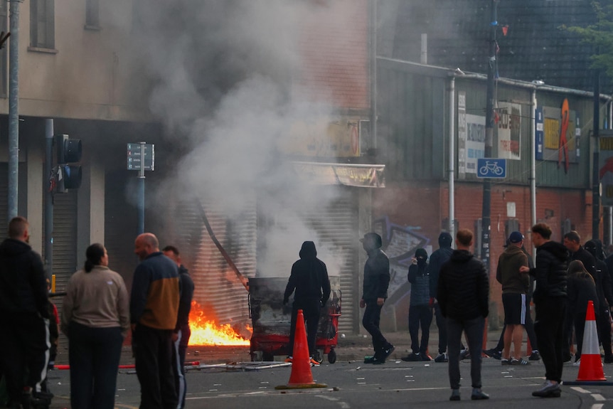 A European building smoulders after civil unrest as people walk restlessly on the street