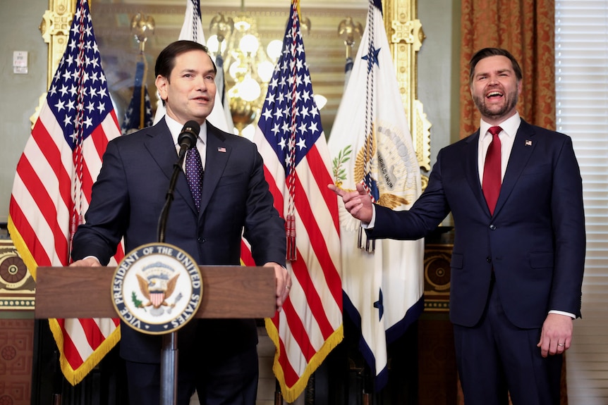 Marco Rubio speaks at a podium as JD Vance stands beside him, pointing and laughing.